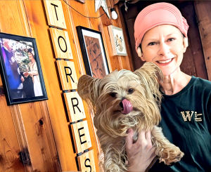 Northwest New Jersey local Kimberly Casteel Torres holding her Yorkie Pablo, standing in front of TORRES wall tiles and a wedding photo, wearing a Women at Green Thumb shirt.